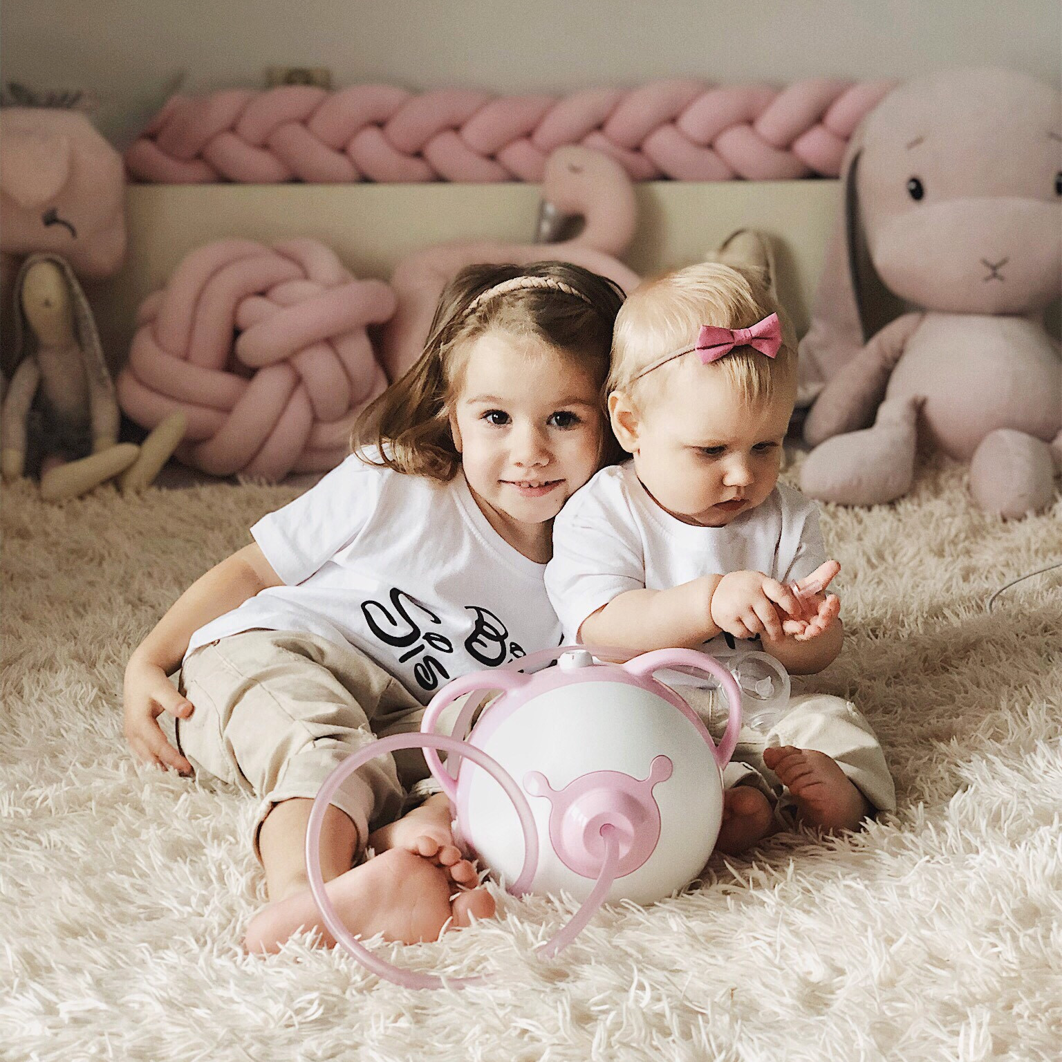Két aranyos kislány ülnek az ágyon, előttük egy pink Nosiboo Pro elektromos orrszívó