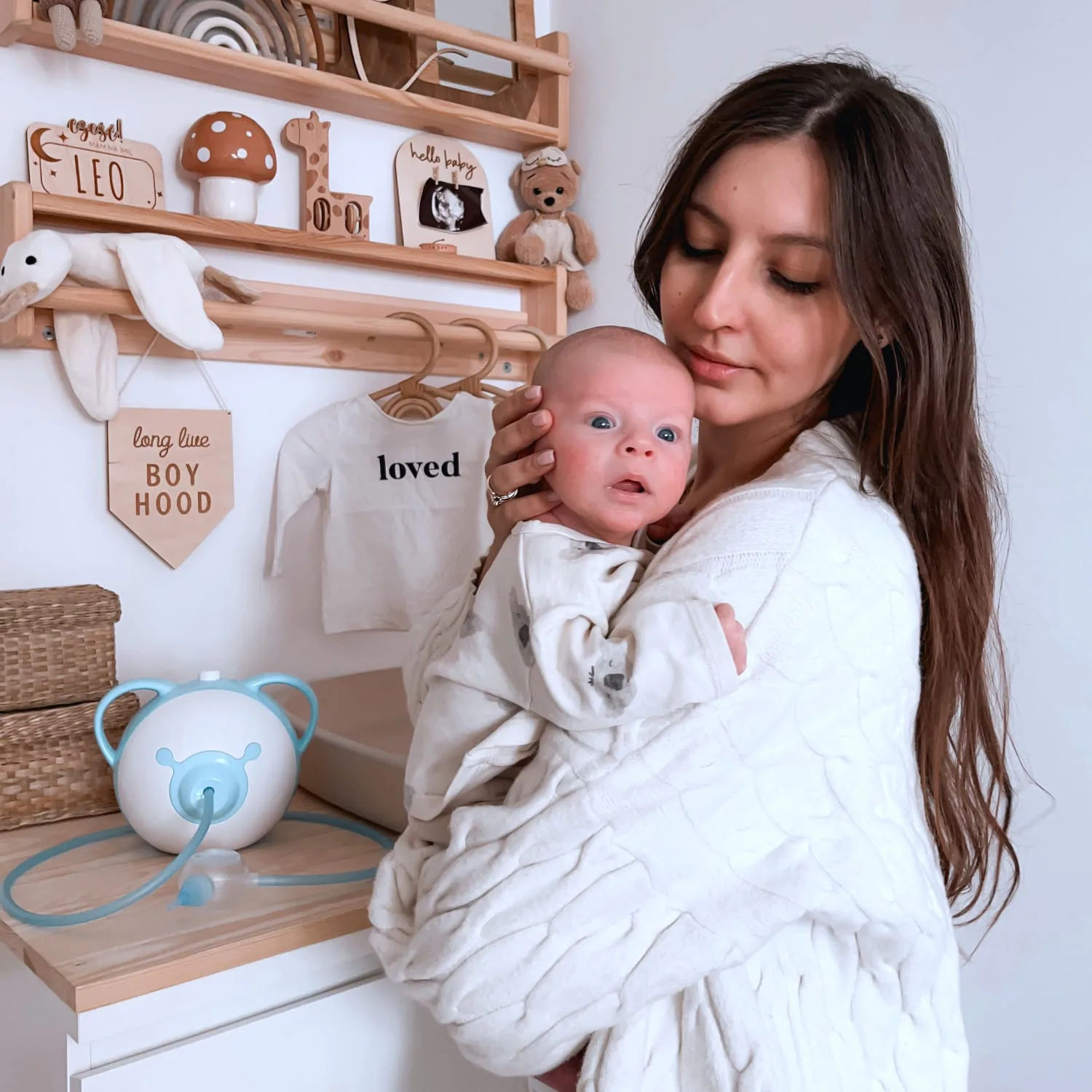 A pretty mother holding her cute newborn baby in her arms, standing in a children's room next to a Nosiboo Pro electric nasal aspirator.